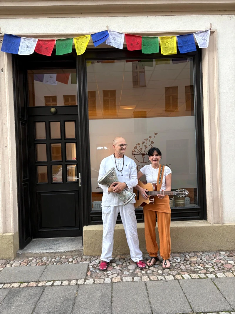 Falk mit Trommel und Jana mit Gitarre vor dem Eingang, darüber bunte Gebetsfahnen.