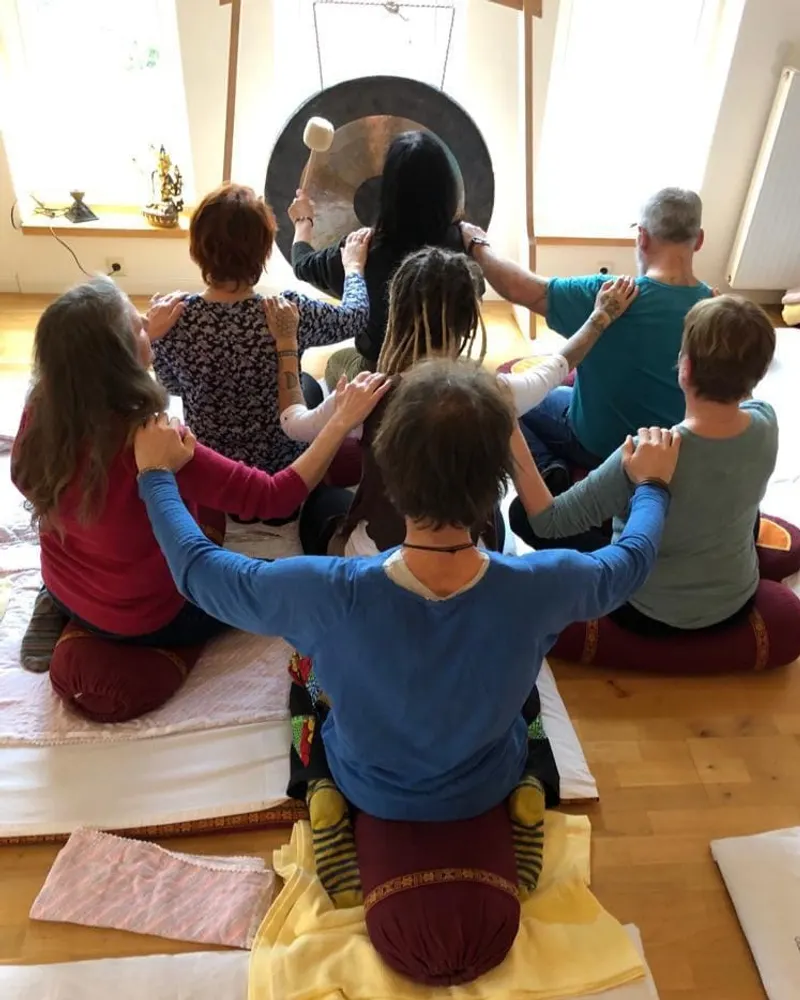Eine Gruppe sitzt im Kreis vor einem Gong, die Hände auf den Schultern der Vordermenschen.
